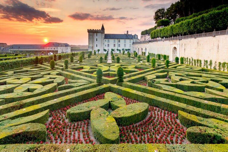 The image shows a formal French garden with meticulously trimmed hedges forming intricate geometric patterns, surrounded by vibrant flowerbeds. In the background, a large historic chateau stands with a stone wall extending along the right side, set against a picturesque sunset sky. This is likely a representation of the gardens at Château de Villandry in the Loire Valley, famous for its beautifully designed Renaissance gardens.