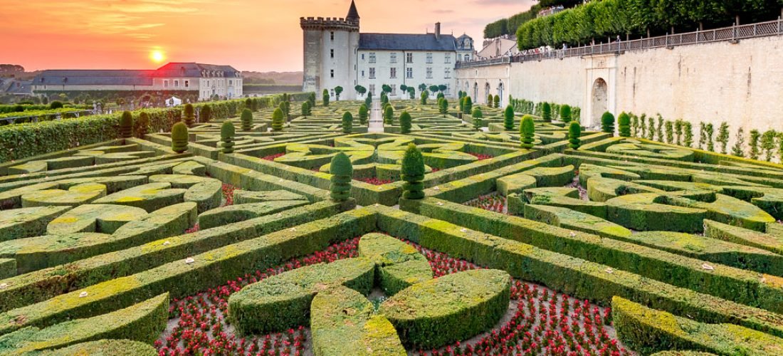 what is loire valley famous for The image shows a formal French garden with meticulously trimmed hedges forming intricate geometric patterns, surrounded by vibrant flowerbeds. In the background, a large historic chateau stands with a stone wall extending along the right side, set against a picturesque sunset sky. This is likely a representation of the gardens at Château de Villandry in the Loire Valley, famous for its beautifully designed Renaissance gardens.